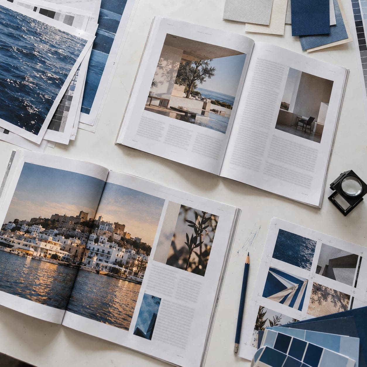 Printed magazine spreads, colour proofs, a loupe and a blue pencil laid out on a white table